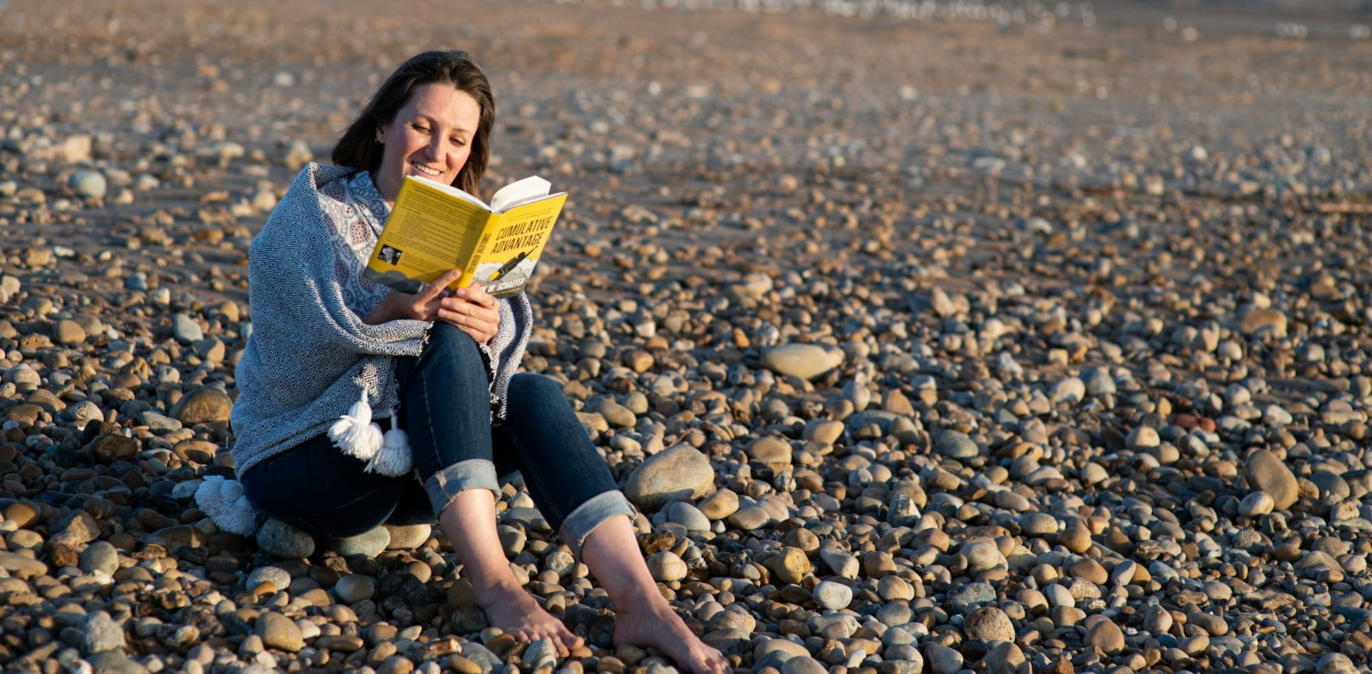 woman in blue denim jacket sitting on rocky shore reading book during daytime